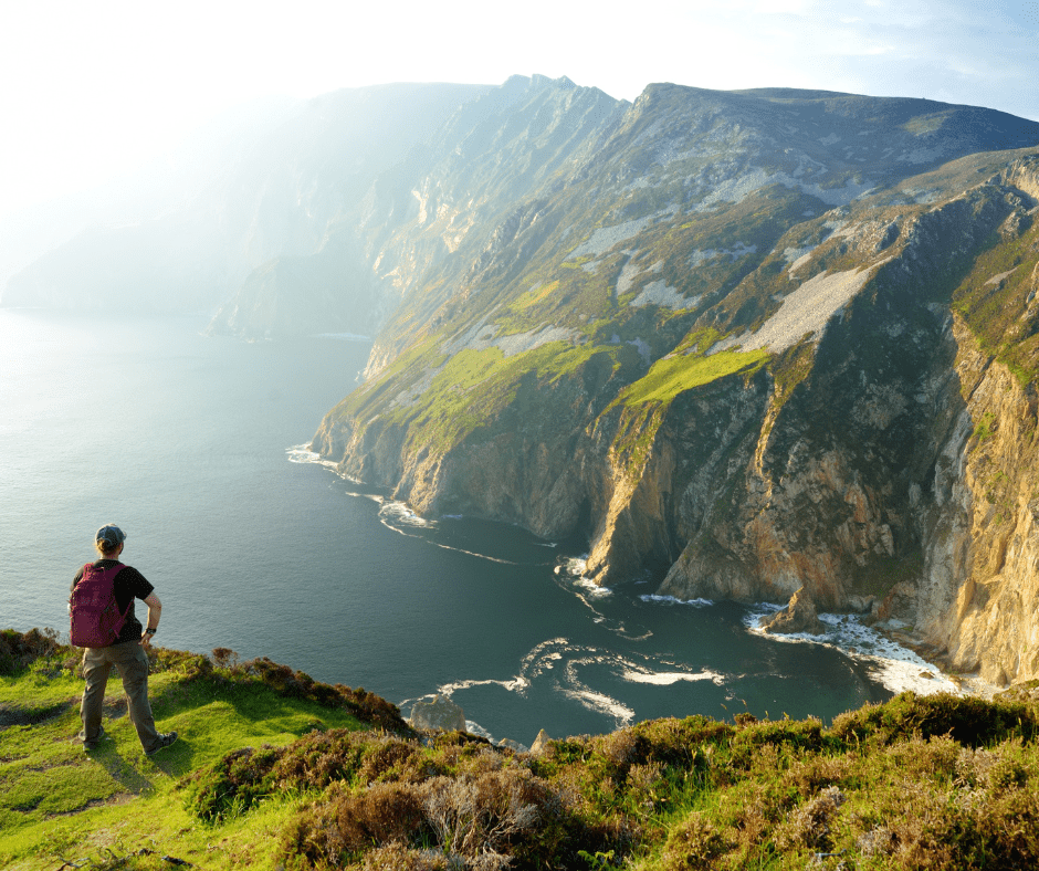 A lone traveler standing near the edge of Slieve League, Ireland’s highest sea cliffs, overlooking dramatic Atlantic views and rugged coastal cliffs under a bright sky.