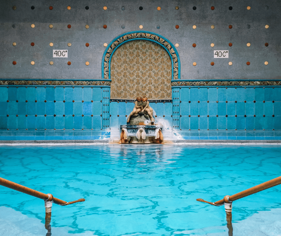 Ornate indoor thermal pool at Gellért Spa in Budapest, Hungary, featuring bright blue water, mosaic-tiled columns, and elegant Art Nouveau architecture.