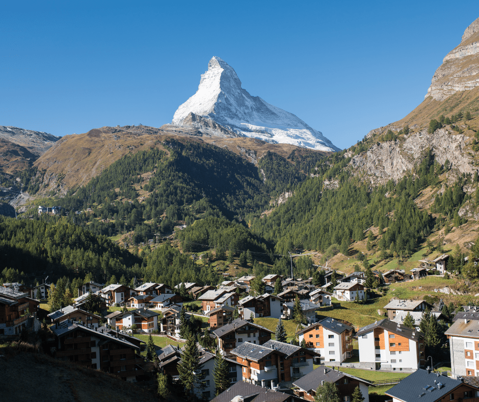 Breathtaking view of Zermatt, Switzerland, with the iconic Matterhorn peak towering over the charming car-free village, surrounded by lush alpine meadows, crisp blue skies, and snow-covered mountains, offering a serene and picturesque Swiss alpine landscape.