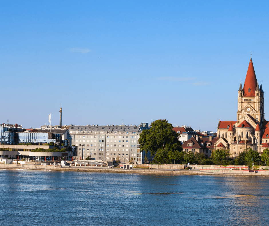 The Danube River flowing through Vienna, Austria, with a cityscape view of historic architecture, church spires, and riverside greenery under a clear sky.