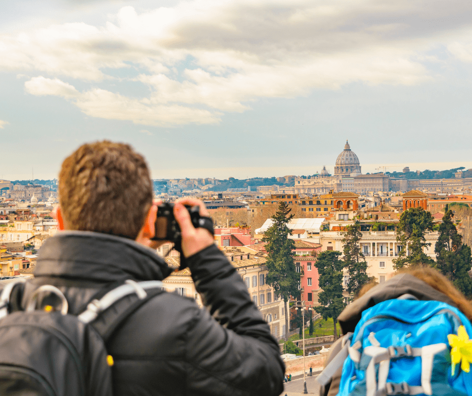 Tourist taking a photo of the Rome cityscape at sunrise, with warm sunlight illuminating rooftops and historic buildings in the distance.